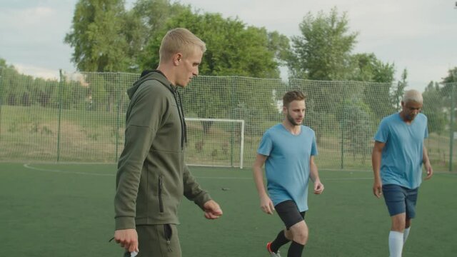 Young football coach using whistle to start performing of hip flexor stretching exercise by team of diverse multiethnic soccer players in sportwear outdoors at daybreak. Male sportsmen stretching legs