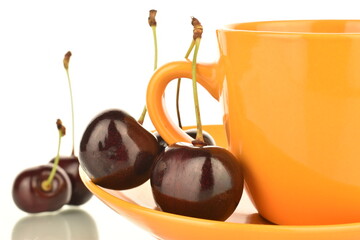 Several juicy berries of dark red cherries on a yellow saucer with a ceramic cup, close-up, isolated on white.