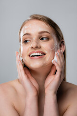 joyful young woman with blue eyes and cleansing foam on face isolated on grey