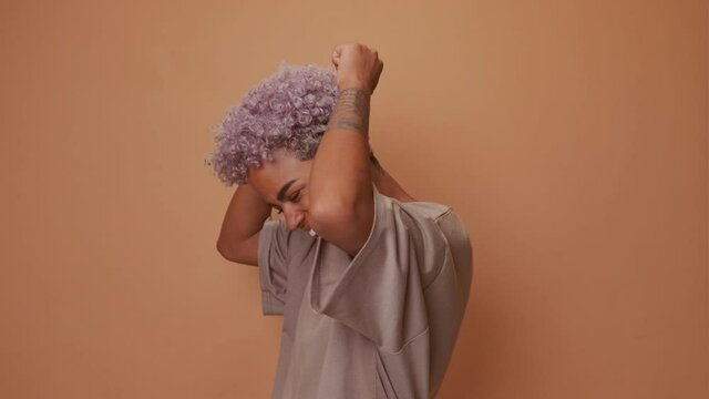 Excited joyous young cute African American woman raises clenched fists, stands without make up against brown background, has curly bushy hair celebrates victory and triumph wears beige t shirt. Oh yes