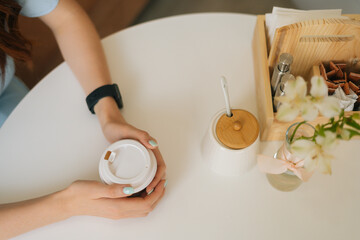 High-angle view of unrecognizable woman holding cup of morning hot coffee sitting at white table in cozy cafe. Top view of beautiful redhead female dreaming with cup of hot cappuccino.