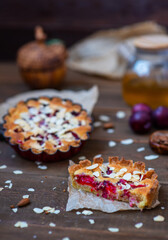 Sandy plum tarts with frangipan and almond petals on a wooden background decorated with plums, one cake is cut and the other is on the background.