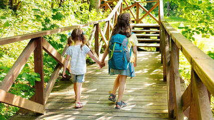 Children's tourism concept. Kids with backpacks go hiking in the reserve. The two sisters head to the family camp along a beautiful hiking trail in a mountain forest.