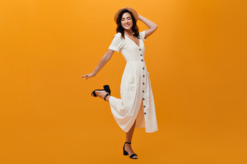 Cheerful girl in mini dress and hat coquettishly raises her leg on orange background