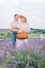 Mature loving couple in the lavender field, standing and hugging, looking each other. Woman in dress and hat holds wicker bag for harvesting lavender