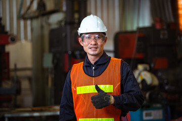 Portrait of Asian male worker in protection glasses and helmet smile showing thumbs up in front of industry machine at industrial factory. quality control guarantee with copy space