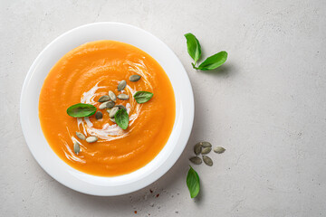 Pumpkin soup puree with cream and basil in a white plate on a gray background.