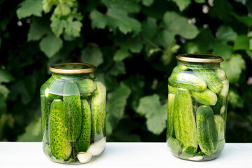 Two cans of canned cucumbers and garlic stand on a white table against the background of green grape leaves