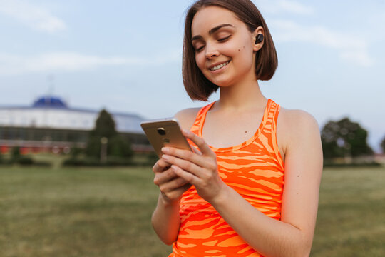 Smiling fit sportswoman in wireless earbuds listening to music during training in city and using smartphone 