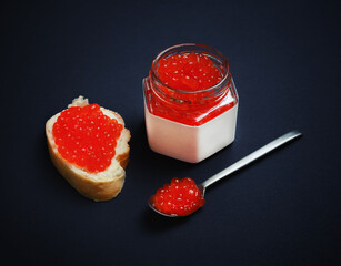 Still life with red caviar. Fresh caviar, sandwich, glass jar and spoon on black background.