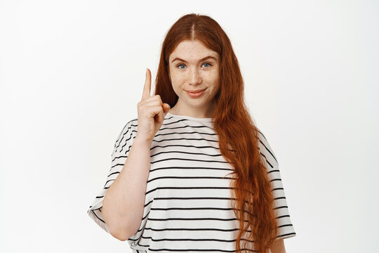 Image of confident young woman with long red hair and freckles, pointing finger up, showing advertisement above, top sale banner, standing against white background