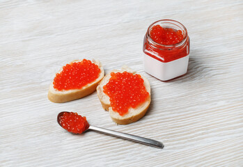 Still life with red caviar. Sandwiches with red caviar, glass jar and spoon on light wood table background.