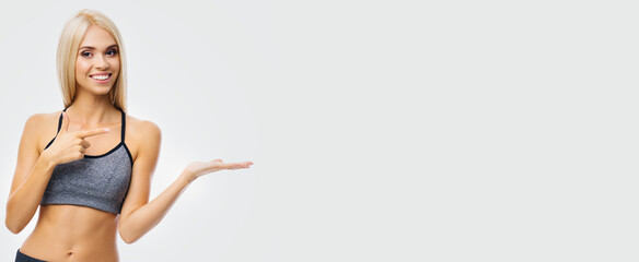 Beautiful smiling woman in sportswear showing, advertising, holding or giving some product or copy space area for text, over grey. Blond girl in fitness studio concept. Wide banner composition image.