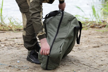 A fisher man packs an outboard boat motor in a carrying bag outdoor on sandy shore grass and water...
