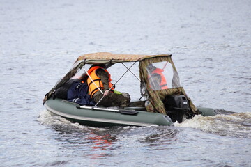 Top awning inflatable green motor boat with two fisherman people in yellow lifejackets with outboard motor floats on the water at autun day, watercraft travel outdoor