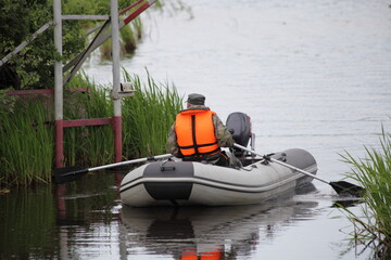 A fisher man in orange lifejacket paddles on an gray inflatable motor boat with broken outboard motor on the calm water, returning to the fishing base entrance gate after fishing at summer morning