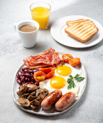 English Breakfast with fried eggs, sausages, bacon, beans, toasts, tomatoes and mushrooms on white plate, concrette background, Top view
