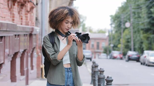 Stylish attractive curly African American black woman walking street and exploring sightseeing with camera. Young female traveler backpacker taking photos against background of blurred brick building.