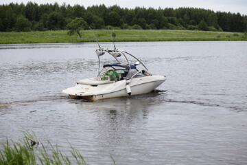 White powerboat with towing targa slowly float on river calm water on green grassy shore with forest background at summer day, front view, active outdoor recreation watersports in Europe