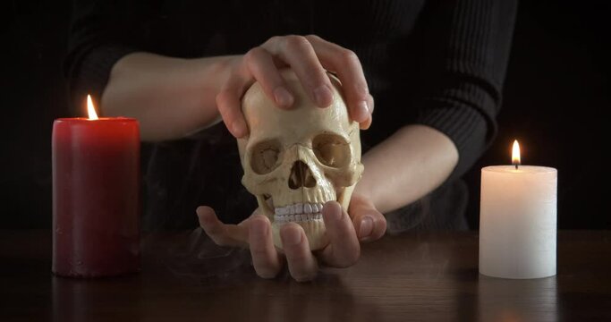 Woman and occultism. A view of a woman making an occultism ritual with a human skull by the table in the dark.