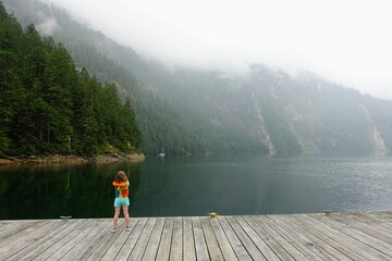 A little girl with her life jacket on standing on a dock admiring the view of the misty princess louisa inlet and surrounding forest, in British Columbia, Canada.