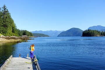 Fototapeta na wymiar A little girl standing on a dock wearing her life jacket with a net looking to catch fishes, with a beautiful background of calm blue ocean and mountains covered in forests in Egmont, British Columbia