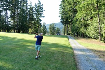 An older male swinging his club from the rough on an approach shot on a par 4 surrounded by forest,...