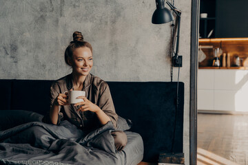 Joyful happy smiling caucasian female in satin cozy pajama holding cup of coffee