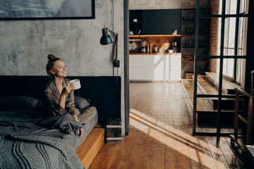 Young happy attractive girl sitting on bed in lotus position and drinking morning coffee