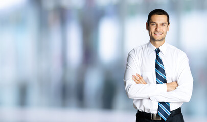 Portrait of happy smiling businessman in white shirt and tie, in crossed arm pose, over blurred modern office interior background. Confident business man. Copy space area.