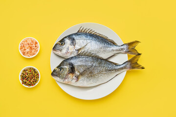 Raw dorado fish with spices on a white plate on a yellow background. Flat lay