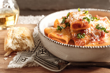 Inviting close-up of a pasta bowl with large rigatoni noodles in a tomato or arrabbiata sauce topped with parmesan and shredded basil.  Bowl sits on a patterned global looking napkin with torn bread. 