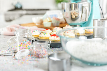 Obraz na płótnie Canvas Pastel kitchen scene with cupcakes. Teal mixer sits with a cupcake baking tray full of cupcakes decorated with red and multicolor sprinkles. Foreground has sugar and flour and jars with sprinkles.
