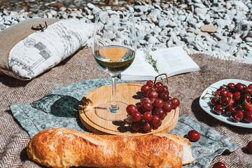 Summer romantic rustic picnic on the coast with white wine glass, baguette, cherries, grape, open book, pillow. Sea beach. Vacation, rest in Europe. Alcohol drink. 