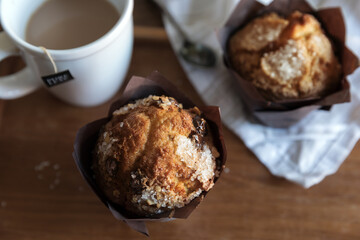 Overhead shot of two sugared chocolate chip muffins with a mug of tea.  Warm, cozy breakfast feel to the shot. Muffins sit on top of a wood surface, one muffin is on a linen napkin with a spoon.  