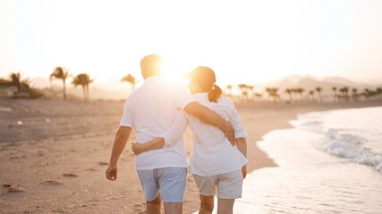 Happy mature senior couple walking and looking at each other on beach during sunset. Aging together and retirement lifestyle concept                               