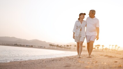 Happy mature senior couple walking and looking at each other on beach during sunset. Aging together...