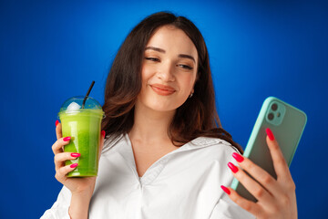 Young attractive happy woman holding a cup of green smoothie and using her smartphone against blue...