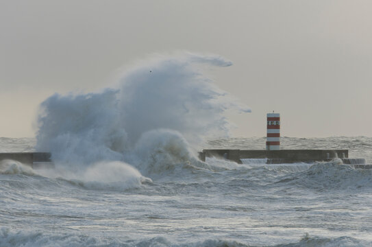 imagen de una ola impactando contra un rompeolas con el faro al lado