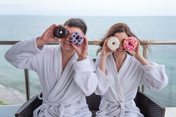 Young couple in love wearing white bathrobe have fun with bright donuts applying to the eyes as glasses. Man woman laugh on the background of the blue sea on vacation travel. Newlyweds honeymoon