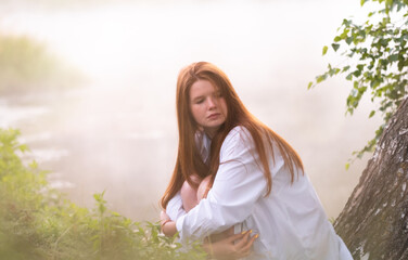 Fototapeta na wymiar A beautiful red-haired girl in a white shirt sits by the lake on a tree in the morning fog. Meeting the dawn in nature.