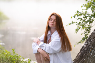 Fototapeta premium A beautiful red-haired girl in a white shirt sits by the lake on a tree in the morning fog. Meeting the dawn in nature.