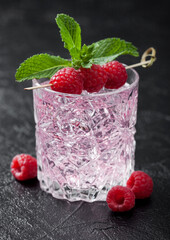 Glass of summer pink lemonade cocktail with raspberries, mint and ice cubes on black background.