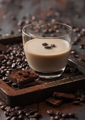 Irish cream baileys liqueur in glass in wooden tray with coffee beans and powder with dark chocolate on dark wood background.