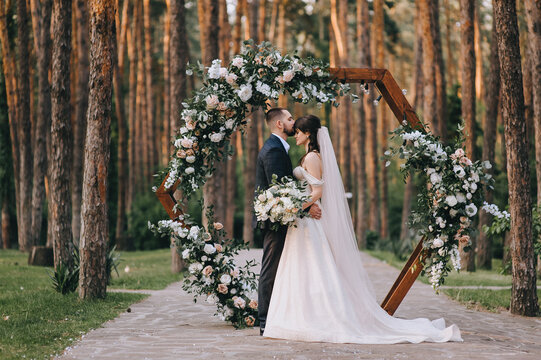Stylish groom in a suit and a cute brunette bride in a white dress in the forest near a wedding wooden arch decorated with flowers. Wedding portrait of the newlyweds.
