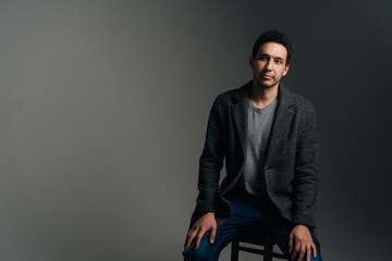 Fototapeta premium Portrait of confident young man wearing stylish long coat sitting in chair on black isolated background looking at camera. Studio shot of serious Caucasian male posing against dark wall.