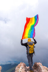 A hiker with a backpack standing on the top of the mountain and holding a pride flag