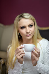 Beautiful blonde girl in a white dress holding a white cup of tea or coffee and sits in a restaurant. Vertical photo