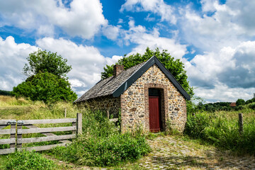 Fototapeta na wymiar Artistic summer rural landscape with an old stone farmhouse and a green field against a bright blue sky. Landmarks and villages in the hilly Dutch Limburg.
