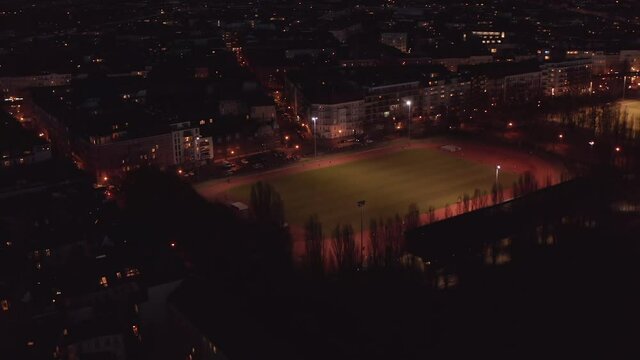 Aerial view of athletes running on oval track. Tilt down footage of artificially lit sports area in late evening. Berlin, Germany.